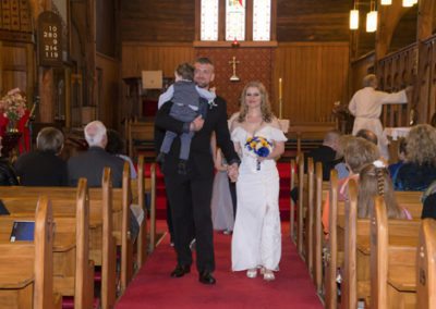 smiling bride & groom walking hand in hand down aisle with groom carrying toddler son