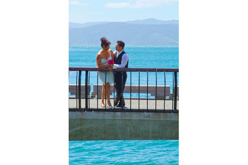 Windswept bride & groom leaning on handrail of waterfront bridge, over beautiful blue water