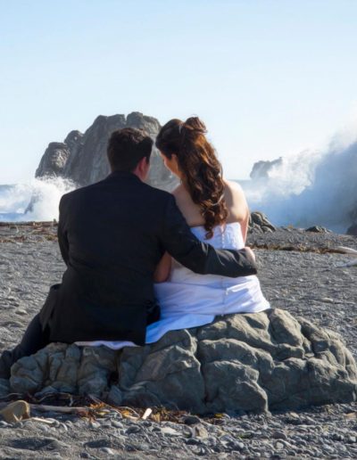 Pencarrow Lodge wedding bride & groom sit on rocks on beach watching large surf