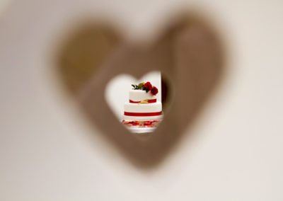 wedding cake with red ribbons and flowers viewed through heart shaped hole in fence