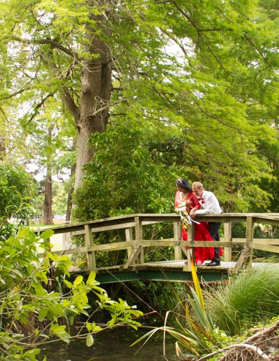 Brightly dressed bride & groom leaning on sides of rustic wooden bridge, in a beautiful bush setting