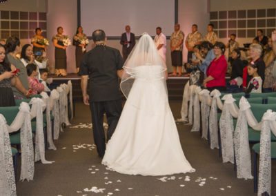 Bride & father walking down lace-bow lined, petal strewn aisle, with groom, bridal party and celebrant waiting on stage