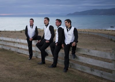 Pencarrow Lodge wedding groom & groomsmen sitting on rustic wooden fence at sunset, with Wellington hills in background