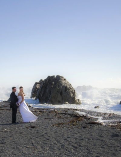 Pencarrow Lodge wedding windswept bride & groom stand on beach with huge surf breaking