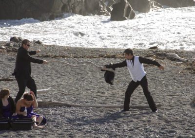 Pencarrow Lodge wedding groomsmen play with driftwood swords on beach as bridesmaids watch from picnic blanket