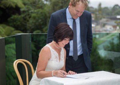 smiling bride signing register outdoors, with groom watching on