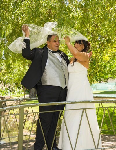 Maori bride & groom laugh as they wrestle with windswept veil on bridge under willow branches