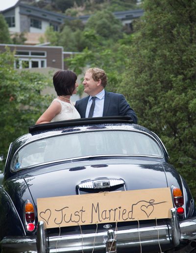 bride & groom standing up through Daimler sunroof, smiling happily at each other, and Just married sign on back of car