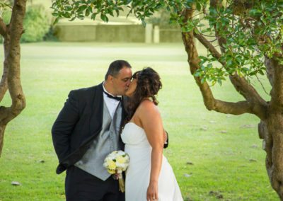 Maori bride & groom kiss in park framed by leafy gnarled trees