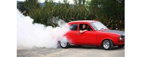smiling young man spinning wheels & throwing up clouds of dirt in red Ford Escort