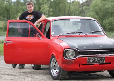 smiling young man leaning against open door of red Ford Escort