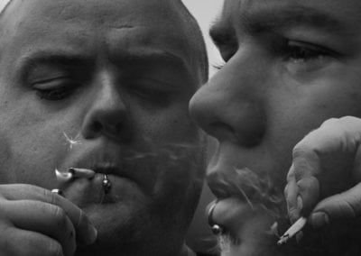 Black & white, man with shaved head & piercings lighting a cigarette