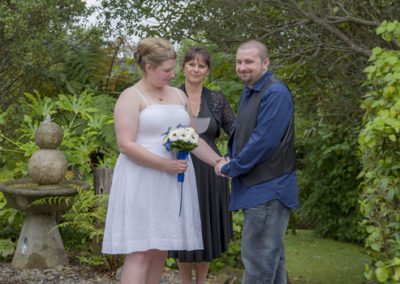 smiling bride & groom holding hands in garden with celebrant looking on