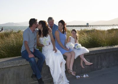 bridal party on Petone beach wall, bride & groom kissing, flower girl giggling