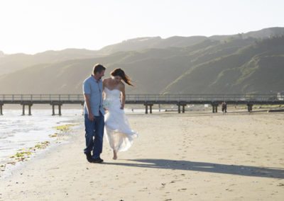 windswept bride & groom walking arm in arm along beach with long sunset shadows