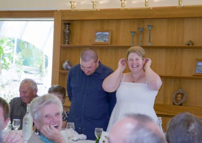 Bridal couple making speeches, laughing bride with her hands raised, and crowd laughing