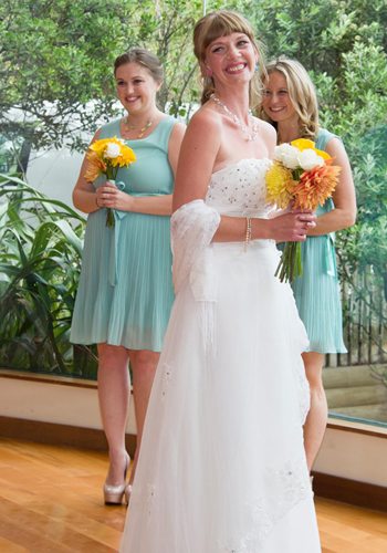 radiant bride holding colourful bouquet and smiling bridesmaids in background, beautiful bushy setting