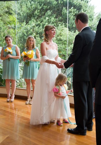 bride & groom holding hands & little girl playing with ribbon, beautiful bushy setting