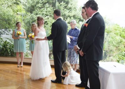 bride & groom holding hands while little girl plays around groom's legs, beautiful bushy setting