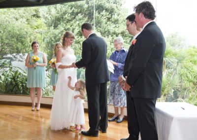 bride & groom exchanging rings with little girl reaching up and laughing, beautiful bushy setting