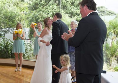 first kiss with happy wedding party clapping in background & smiling celebrant, beautiful bushy setting