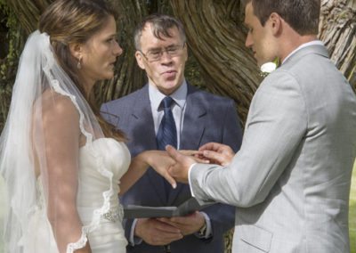 Groom putting ring on bride's finger with smiling celebrant behind them