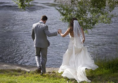 Groom holds windswept bride's hand as he leads her to riverbank