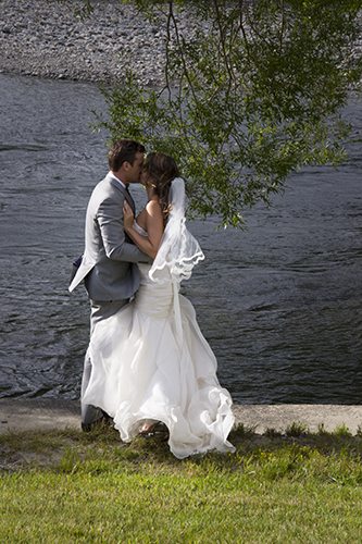Windswept bride and groom share kiss on riverbank