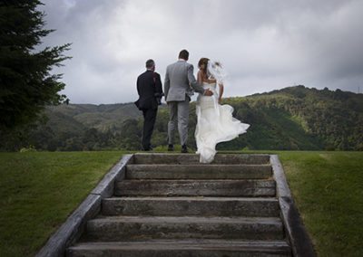 Under a stormy sky, windswept bride & groom walking up steps with groom's father