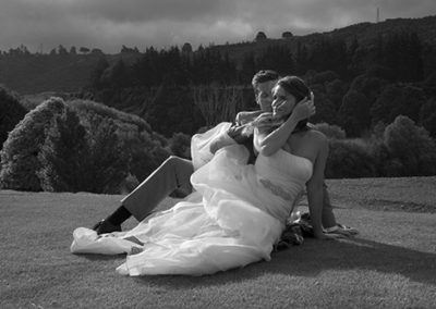 Black & white, windblown bride & groom sitting on grass with bride sweeping hair out of her eyes